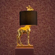 Indlæs billede til gallerivisning Lucie Giraffe Table Lamp in gold with black shade on top of pedestal with leaf wallpaper background